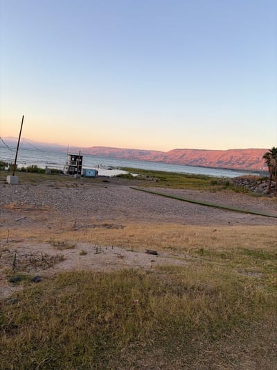 The image depicts a tranquil scene at sunset near the Sea of Galilee, within the Emek HaYarden Regional Council, Israel. The sky transitions from a clear pale blue at the top to a warm, soft orange hue near the horizon, indicating the setting sun. The Sea of Galilee stretches across the middle ground, its surface calm and reflecting the colors of the sky. Across the water, a range of hills is illuminated by the golden light of sunset, their contours sharply defined against the sky.

In the foreground and middle ground, the landscape is a mixture of dry, golden-brown grass and coarse gravel. A prominent structure, appearing to be a small, multi-story building with windows and a dark roof, stands on the rocky shoreline. Adjacent to it is a light blue cylindrical tank with branding that is not clearly legible. To the left of the building, a tall, slender pole with a concrete base stands firmly planted. A narrow, dark green strip of material, possibly a pathway or mat, runs diagonally across the gravelly area.

On the right side of the image, a tall palm tree stands against the rocky embankment. Several people are visible near the water's edge, some appearing to be wading or standing in the shallow water. The overall impression is one of peace and natural beauty, with the human elements integrated into the serene landscape.