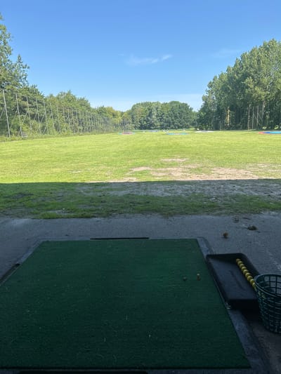 The image shows an outdoor golf driving range on a clear, sunny day. In the foreground, a green artificial turf mat is positioned on a concrete surface, with two small wooden tee holders visible. To the right of the mat, a black tray holds a row of yellow golf balls, and a green plastic basket is nearby. The driving range itself is a large, grassy field with various colored targets scattered across it at different distances. The field is bordered by trees and a tall fence. The scene appears to be located at the Golfclub Wassenaar, situated in the municipality of Wassenaar, Netherlands, given the presence of a golf course. No people are visible in the image.