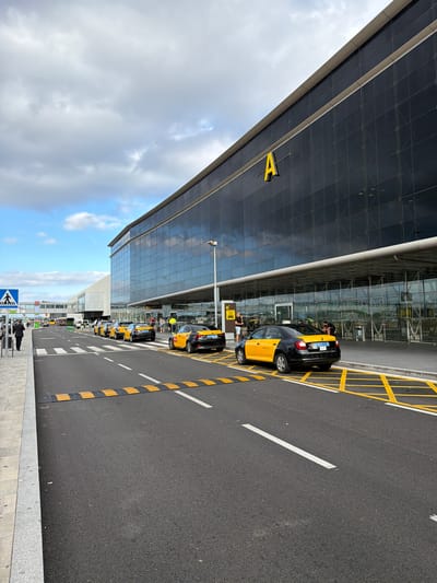 The image shows the exterior of Terminal A at Barcelona-El Prat Airport in El Prat de Llobregat, Spain. A line of black and yellow taxis is parked on the roadway in front of the terminal's glass facade. The terminal building features large, dark windows and a prominent yellow letter "A" on its upper level. A pedestrian crossing with a blue sign is visible on the left side of the road. Several people are present, including a person walking on the sidewalk, individuals standing near the taxis, and others near the entrance of the terminal. The sky is partly cloudy with patches of blue. Yellow and white road markings, including a speed bump, are present on the asphalt.
