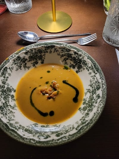 A bowl of orange soup, decorated to resemble a smiley face with green oil, sits on a floral-patterned plate. Scattered toppings rest in the center. A spoon and fork lie beside a gold coaster. A partially visible glass is near the top-left, and another glass sits near the bottom-left of the dark wood table.