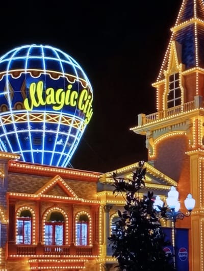 The image depicts a nighttime scene in Aktash, Uzbekistan, focused on an illuminated entertainment complex. A large, blue hot air balloon-shaped structure, outlined with bright blue lights and bearing the yellow text "Magic City" on its side, dominates the upper left. Below and to the right, multiple buildings are extensively decorated with numerous small, warm white lights outlining their architectural features, including windows, gables, and cornices. On the far right, a tall, multi-tiered tower-like structure with a blue roof is similarly adorned with warm white lights. In the lower right foreground, a dark-leaved tree partially obscures a two-headed white streetlamp and a blue banner displaying the Pepsi logo. The sky is dark, indicating a nighttime setting. No individuals or active subjects are present.