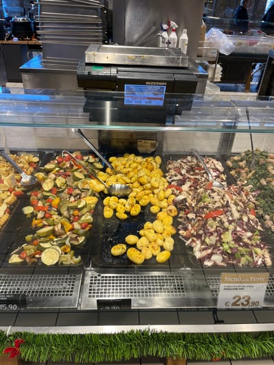 The media file captures an indoor food display counter, likely within a supermarket or deli in Rozzano, Italy. Various prepared food items are showcased in transparent containers behind a glass barrier, suggesting a self-service or assisted-service buffet.
The display features four distinct dishes:
* The topmost container, partially visible, appears to hold a mixed greens or vegetable salad.
* Below it, a full container holds "Insalata di pollo alla senape" (chicken salad with mustard), containing chopped chicken, radicchio, and other ingredients. This dish is priced at €23.90, as indicated by a sign also reading "Menú delle Feste" (Holiday Menu) and "PRODUZIONE IPER".
* The middle container is filled with golden-brown roasted potatoes, seasoned with herbs like rosemary. A partially visible sign indicates "Patate al Forno" (Baked Potatoes) for €15.90.
* The bottom container contains mixed roasted vegetables, including zucchini, bell peppers, and possibly onions, also priced at €15.90 for "Miste" (mixed).
Serving spoons are placed in the potato and mixed vegetable containers. To the left of the food display, a Bizerba brand weighing scale is visible. Its digital display shows "0.020 0.020 34.90", indicating a weight of 0.020 units and a price of €34.90, likely per kilogram. The background shows stainless steel surfaces, suggesting a commercial kitchen or food preparation area. A green, artificial garland runs along the right side of the display, hinting at a festive or holiday season.