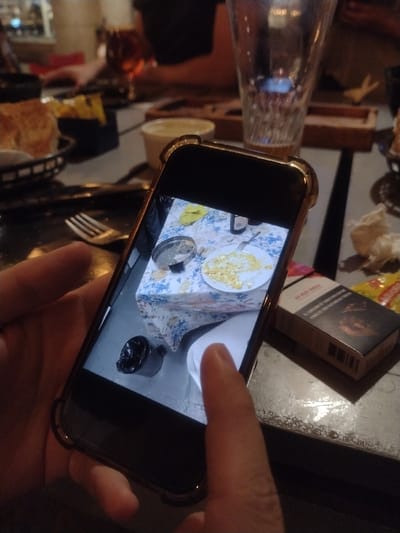 A hand holds a smartphone displaying a photo of a breakfast table setting. Scrambled eggs on a plate, a side of yellow fruit, silverware, a white napkin, and a small black cup are captured on the phone screen. In the background, there is a partially visible cigarette pack, a glass carafe, and what appears to be other food and restaurant items on a table. The image suggests someone taking a picture of their meal.