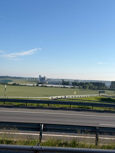 The image shows a landscape with a highway in the foreground and agricultural fields and industrial buildings in the background. The sky is clear and blue. The highway has guardrails and white lines marking the lanes. The fields are green and appear to be planted with crops. In the distance, there are several industrial buildings, including a silo and a long, low structure with a green roof. There are also trees and other vegetation. The overall scene is rural and appears to be taken on a sunny day. The location is identified as being in the city of Borskoe, Russia. There are no people or animals visible in the image.