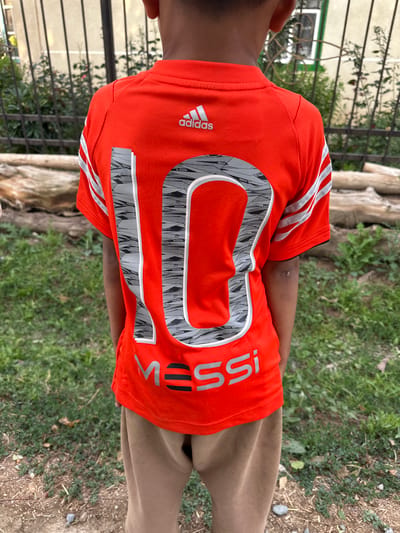 The media file features a young person wearing an orange Adidas t-shirt with a large "10" printed on the back, along with the name "MESSI." The shirt has the signature Adidas stripes on the sleeves. The subject is standing outdoors, as indicated by the presence of grass, dirt, and what appears to be a tree trunk in the background. There is also a metal fence visible on the left side of the image. The weather appears to be overcast or diffused daylight, as there are no strong shadows. The setting suggests a casual outdoor environment, potentially a backyard or a park area. The person's arm is visible, and they are wearing brown pants. The overall tone is casual and sporty. The visible text includes "adidas," "MESSI," and the number "10."