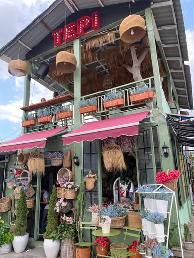 A two-story green building with a corrugated metal roof is pictured on a cloudy day. The building is decorated with numerous hanging woven baskets and planters filled with flowers and greenery. The words "TEPI" in red, illuminated lettering are visible on the upper story. The lower story features a red awning over large windows, and the entrance is adorned with a collection of various woven baskets and plants. A person wearing a gray shirt and dark pants can be seen inside the doorway. The scene is consistent with a cafe or shop in Pasar Pangururan, Indonesia, given its architectural style and the presence of outdoor seating and decorative elements.
