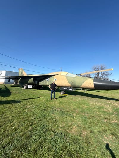 A camouflaged jet sits on a grassy field under a clear blue sky. A man in a black shirt, jeans, and a cowboy hat stands beside the aircraft, posing with his hands on his hips. A small white building with a dark roof is visible in the lower left corner of the frame. Overhead power lines are strung across the sky. A bare tree stands behind the jet.