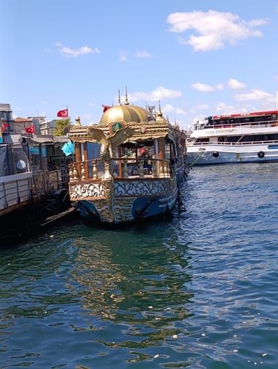 The image depicts a sunny day in Fatih, Turkey, with a view of the Golden Horn waterway. In the foreground, a highly ornate, gold-trimmed boat with a domed roof and eagle motifs is docked. The boat has intricate carvings and a railing along its deck. To the left of this boat, another, more utilitarian vessel is moored. In the background, a larger white ferry with red accents is visible, also on the water. The sky is blue with scattered white clouds. Several Turkish flags are flying from the boats and buildings on the shore. The water is a deep blue-green, reflecting the sunlight and the boats. People are visible on the decks of the boats, and buildings line the distant shore, suggesting an urban waterfront.