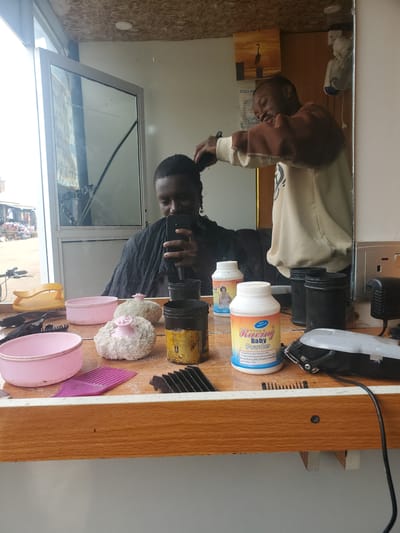 In a barbershop in Ohit, Nigeria, a barber reflected in a mirror is cutting the hair of a client. The client is seated and wearing a black cape, holding a smartphone to take a selfie. The barber, wearing a brown and beige hooded sweatshirt, is using clippers to cut the client's hair. On the wooden counter in front of them, various barber tools are visible, including pink bowls, a yellow hair dryer, combs, and containers of talcum powder. A hair clipper is also on the counter, plugged in and ready for use. The shop appears to be dimly lit, with natural light filtering in from a large window. The background shows a glimpse of the bustling city street outside.