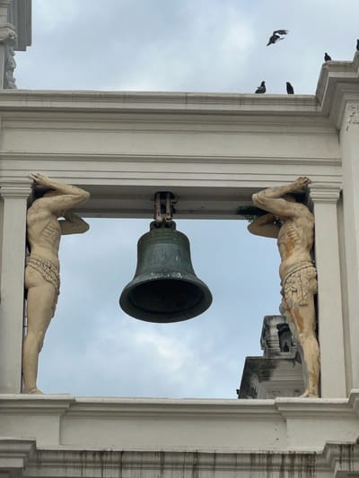 This image shows a view looking up at a large bell suspended between two architectural statues, located on the facade of the León Cathedral in León, Nicaragua. The statues depict male figures with their arms raised, appearing to support the structure above. The bell is a dark, metallic color. Several pigeons are perched on the architectural elements and in the sky above. The sky is overcast and a light blue-grey. The overall scene is one of architectural detail and avian presence against a cloudy sky.