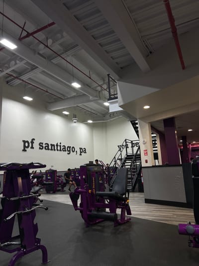 This image depicts the interior of a Planet Fitness gymnasium located in Nuevo Santiago, Panama. The scene is brightly illuminated by overhead rectangular fluorescent lights, set against a high, industrial-style ceiling featuring exposed white corrugated metal panels, large white cylindrical air ducts, and red fire suppression pipes. A white security camera is mounted near the ceiling in the background.
The left-most wall is white, displaying "pf santiago, pa" in large, black, sans-serif lettering. The gym floor features two-tone gray rubberized matting, transitioning from a darker shade in the foreground to a lighter, possibly patterned, gray in the mid-ground and right.
Numerous purple and black exercise machines, characteristic of Planet Fitness, are arranged throughout the space. In the mid-ground, a male individual is seated on a purple and black leg exercise machine, facing away from the viewer, wearing dark attire. Another male is partially visible further back near additional purple equipment. To the right, a black metal staircase with a matching railing ascends to an upper level. Two indistinct figures are visible on this upper level. A prominent purple structural column and a gray, textured counter or partition are also present on the right side of the image. The environment is well-maintained and appears operational.