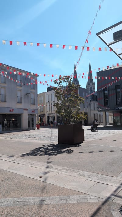 The image shows a sunny day in a pedestrianized shopping area, likely in Cholet, France, given the context of the H&M store. Red and white pennant banners are strung across the sky. On the left, the H&M store is visible with mannequins in the window. Across the street, a U-shaped sign that reads "Arcades Rouge" is prominent. In the background, the spires of a cathedral can be seen. A large tree in a planter is positioned in the middle of the square. Several people are present, including a couple walking, and a person with a stroller. The ground is paved with different textures and patterns. The architecture suggests a European city.