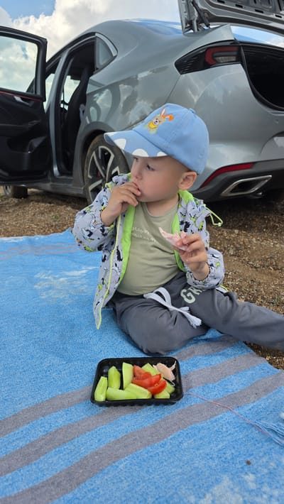 A young child wearing a blue cap with a rabbit design and a grey jacket with a dinosaur pattern is sitting on a blue and grey striped blanket. The child is holding a piece of food, possibly meat, in their right hand and has their left hand near their mouth, suggesting they are eating. In front of the child is a black plastic container with sliced cucumbers and tomatoes. A grey car with its doors and trunk open is parked behind the child. The ground is covered with gravel and dirt. The sky above is cloudy. This scene is taking place outdoors, potentially in a rural or semi-rural area given the presence of gravel and the car.