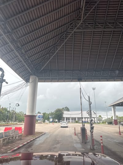 This image depicts an outdoor scene under an overcast sky, likely in the late morning or early afternoon. The perspective is tilted, making it difficult to determine the exact orientation of the scene.
On the left side of the image, the underside of a large, corrugated metal roof is visible, supported by a complex network of metal beams and trusses. This structure appears to be part of a covered outdoor area, possibly a market, stadium, or industrial facility.
The right side of the image shows an outdoor paved area with several objects and structures. These include a large, light-colored cylindrical pillar, a traffic barrier with red and white markings, and what appear to be electrical poles with numerous wires. In the background, there are buildings, trees, and several vehicles, including a white car and the reflection of a dark-colored car in the wet pavement. Red traffic cones and barrier blocks are also present.
There are no people visible in the image. The overall atmosphere is functional, with a focus on infrastructure and utilitarian structures. The wet pavement suggests recent rain or ongoing light precipitation. The location is identified as Ban Tha Chat Chai, Thailand, which aligns with the presence of tropical trees and the style of the buildings and infrastructure. No specific text is legible in the image.