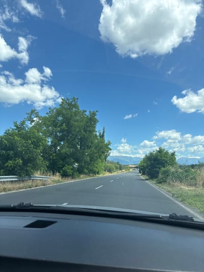 The image captures a view from inside a vehicle driving on a paved road. The road is flanked by lush green trees on the left and right sides, with dry grasses and vegetation also visible. A metal guardrail runs along the left side of the road. In the distance, mountains are visible under a bright blue sky dotted with white, fluffy clouds. The foreground shows the dark dashboard of the vehicle, partially obscuring the view. The scene appears to be in a rural or semi-rural setting, possibly on a highway or main road leading out of or into a town. Based on the information provided about the location, this scene is situated in or near the city of Ivan Vazovo, Bulgaria.