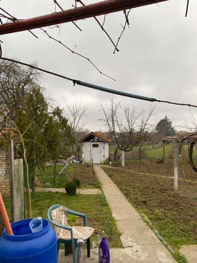 An outdoor scene in Sredkovets, Bulgaria, depicts a residential or garden area under an overcast sky. In the foreground, two horizontal bars, one red-brown and one grey, with bare vine tendrils, span the upper portion of the frame. Below and to the left, a blue plastic barrel contains a white plastic measuring cup and an orange pipe. Adjacent to it, a light green plastic garden chair with a patterned cushion is positioned on a concrete surface. Next to the chair are a clear glass bottle, a grey metallic object, and a purple plastic bottle.

A narrow concrete path extends from the immediate foreground through the midground. To the left of the path, a brick structure is partially visible, followed by an area of patchy grass, a small green shrub, and a tall conifer tree. To the right of the path, the ground consists of disturbed soil and sparse grass, with a bare deciduous tree featuring a white-painted lower trunk.

The path leads to a small, single-story white building constructed of concrete blocks, centrally located in the midground. It has a white door, a small rectangular opening above the door, and a brown-tiled gable roof. Behind this building, additional bare trees are visible, along with a green mesh fence. The background reveals further trees and gently undulating terrain under the consistent overcast conditions. No individuals are present in the image.