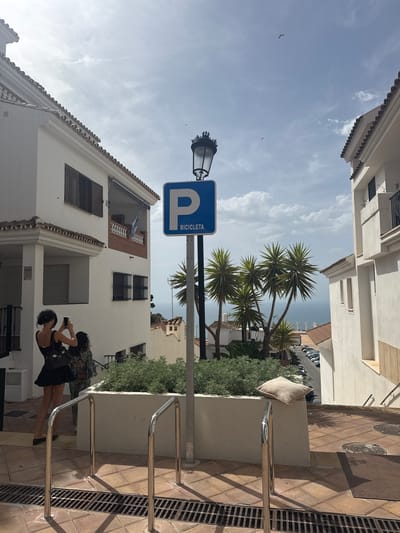 The image shows a street scene in Benalmádena, Spain, with white buildings characteristic of the region. In the foreground, two women are standing near a parking sign for bicycles. One woman, wearing a black dress, is taking a photo with her phone. The other woman is partially obscured. A street lamp with a decorative top is visible above the parking sign. Palm trees are growing in the midground, and beyond them, the Mediterranean Sea can be seen in the distance. A parked car lot is visible down the slope. The scene appears to be on a sunny day, with shadows cast by the buildings and palm trees.