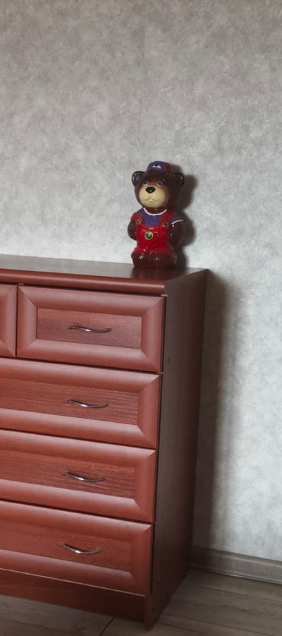 A small, brown ceramic bear figurine with red overalls and a blue hat is standing on the corner of a dark brown wooden dresser. The dresser has several drawers with silver handles. The background is a light gray textured wall. The scene is indoors, possibly in a residential setting. The location is the city of Aktanyshbash, Russia.