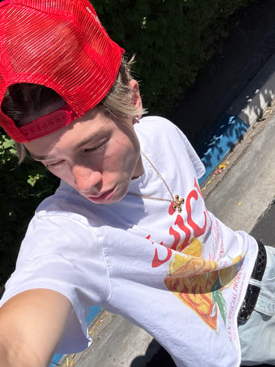 A pale-skinned person is lying down in an outdoor environment, possibly near or in Indian Wells, United States of America. They are wearing a red mesh trucker hat, a white t-shirt with a graphic print that includes the words "MAN OFFICIAL PRODUCT," light denim jeans with a black woven belt, and a gold flower-shaped pendant necklace. The person's eyes are closed, and their face is angled towards the camera. The ground below is covered in patches of green foliage, and a concrete structure or sidewalk with blue markings is visible in the background. The lighting indicates it is daytime, with bright sunlight.