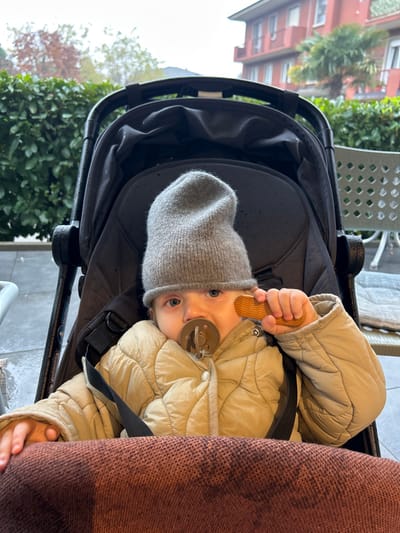 An infant or toddler with fair skin and blue eyes is observed seated in a dark grey or black stroller. The child wears a light grey knit beanie, a light beige quilted puffer jacket, and has a dark brown pacifier in their mouth. Their lower body is covered by a dark reddish-brown knit blanket. The child holds a small, ribbed, light brown wooden object in their right hand. The background reveals an outdoor setting, likely a patio, located in Longone al Segrino, Italy. To the left, a dense green hedge is visible. To the right, a multi-story building with reddish-orange walls and balconies is present, with a green palm tree situated in front of it. Portions of a white metal outdoor chair and table are visible on the far right. The sky appears overcast.
