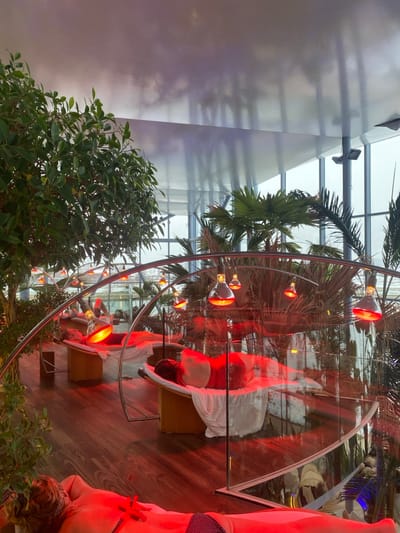 The image captures an indoor thermal spa or wellness area, identified as being in Săftica, Romania. The environment is a large, brightly lit space featuring a high, reflective ceiling and extensive windows that reveal an overcast outdoor setting. The floor is composed of dark wood paneling. Lush green foliage, including tall trees and palm plants, are arranged in large planters throughout the area.

Multiple individuals are present, reclining horizontally on lounge chairs. In the foreground, one individual lies face down, partially clad in dark swimwear with white polka dots, their back illuminated by a prominent red light. Behind them, several other individuals are visible lying supine on similar recliners, partially covered by white towels or sheets, also bathed in red light.

Each recliner is positioned beneath an arched metallic structure from which multiple dome-shaped lamps are suspended. These lamps emit a strong red glow directly onto the individuals below, indicative of infrared heat or light therapy. Curved glass partitions or railings segment portions of the relaxation area.