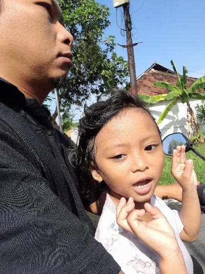 A man in a black shirt holds a child in his arms while on a motorcycle. The child faces the camera, smiling with an open mouth. A motorcycle mirror is visible behind the child. In the background, there's a white building with a brown roof, banana trees, a power pole, and other greenery against a blue sky. The photo is taken from a low angle, slightly rotated.