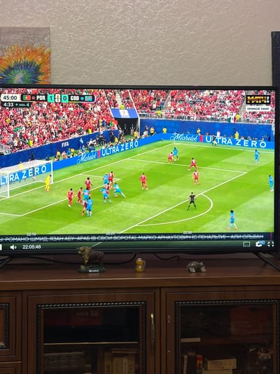 A flat-screen television displays a live broadcast of a soccer match. The score shows Portugal leading 1-0 against COD at 45:00 minutes with 4 minutes of added time. The players are wearing red and blue uniforms. The stadium is filled with spectators. The lower part of the screen shows a graphic overlay with text in Cyrillic, indicating the time 22:05:46. The television is placed on a wooden cabinet. A small figurine of a buffalo with "BELARUS" written underneath it is on the cabinet, along with a small glass object. The background of the image is a textured wallpaper.