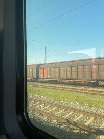 The image presents a railway environment observed from inside a train, looking out a dark gray-framed window. Multiple parallel railway tracks are visible in the foreground and midground, situated on ballast with intermittent patches of green grass. A stationary freight train, composed of several dark brown or olive green boxcars with reddish-brown panels, occupies an adjacent track. One panel displays a white stylized "H" logo, while another shows white numbers including "4324". Overhead power lines are stretched across a clear, bright blue sky, and a metal lattice tower is discernible on the horizon to the left. Sparse vegetation outlines the far background. No individuals or specific actions are visible in the scene, which is under bright daylight. This scene is located in Kalitinovo, Bulgaria.