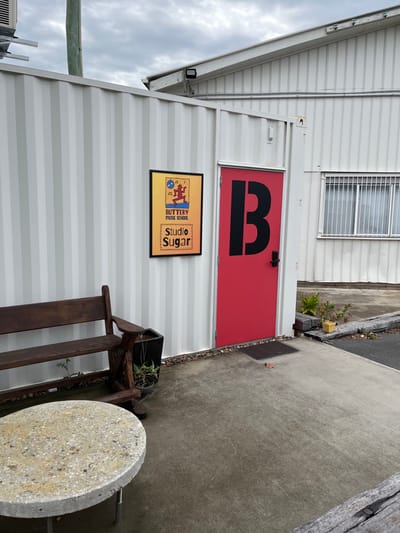 The image displays the exterior of a modern building, likely a modified shipping container or similar modular structure, characterized by its white corrugated metal paneling. The sky is overcast, suggesting daylight under cloudy conditions.

A striking red door, marked with a large black capital "B", serves as a central focal point. To the left of this door, a rectangular sign is mounted on the wall. The sign features a warm yellow-to-orange gradient background and displays the text "BUTTERY MUSIC SCHOOL" vertically alongside musical note symbols, and "Studio Sugar" with a small sugar cube graphic. A stylized figure, possibly dancing or playing an instrument, is also part of the sign's design.

Above the door, a window with horizontal security bars is set into the building's upper section. A concrete path stretches away from the door to the right, with a small dark mat placed directly at the threshold. Along the path's edge, sparse vegetation and a small yellow object are visible.

In the lower right foreground, an outdoor seating arrangement is present, comprising a rustic wooden bench and a round, light-colored table with a speckled, stone-like surface and a single metal leg. A potted plant rests on the wooden bench. No people are visible, and no specific activity is occurring. The scene depicts an entrance area for a music school and studio in Hamilton, Australia.
