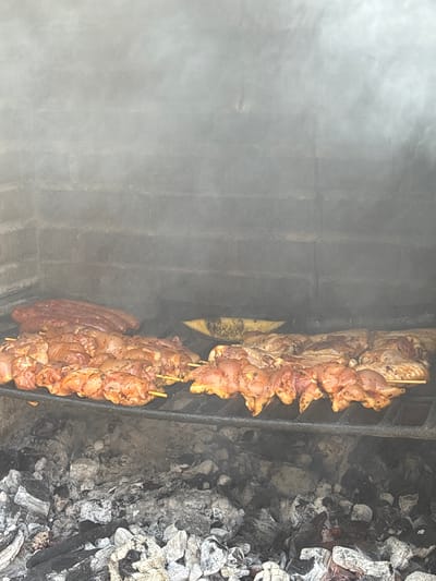 This is a close-up, eye-level, medium shot of a barbecue grill with meat cooking over hot coals in Campezo / Kanpezu, Spain. The grill grate is made of dark metal and is loaded with several rows of skewers holding pieces of seasoned chicken. Behind the chicken skewers, there are several thick sausages on the grill. A hazy smoke fills the upper portion of the image, obscuring the brick interior of the grill. The bottom portion of the image shows a bed of glowing embers and charcoal. There are no people visible in the image.
