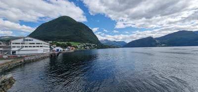 This is a photograph taken from a watercraft, likely a ferry or boat, looking towards the shore of Volda, Norway. The image captures a coastal scene featuring a large body of water in the foreground, reflecting the sky and surrounding landscape. On the left bank, a long, multi-story white building with numerous windows and a red roof is prominent, suggesting it might be a commercial or industrial facility. Adjacent to this building, a rocky shoreline is visible. Further along the coast, a small town or village is nestled at the base of a steep, tree-covered mountain. The town consists of several houses and other structures, painted in various colors, including white, red, and yellow. The mountain rises dramatically behind the town, its slopes densely forested. In the distance, more mountains, some with snow-capped peaks, recede into the background. The sky is partially cloudy, with patches of blue sky interspersed with white clouds. The overall impression is one of a picturesque Norwegian coastal town with a backdrop of imposing natural scenery.