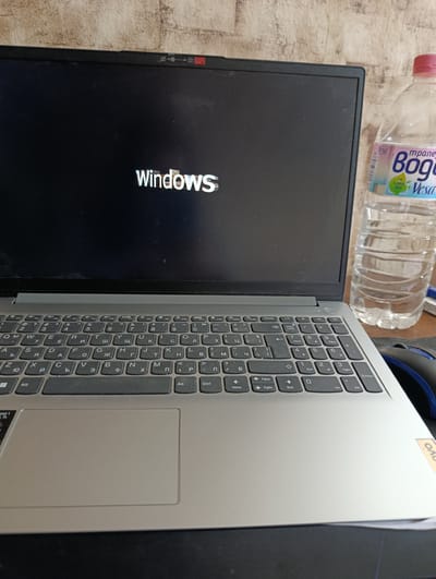 The image shows a laptop computer displaying the Windows logo on a black screen. The laptop appears to be a Lenovo model, indicated by a small sticker visible on the bottom right corner of the base. The keyboard has Cyrillic characters in addition to Latin ones. To the right of the laptop, there is a plastic bottle filled with water; the label on the bottle is partially visible, featuring the word "Vesa" and the word "Bog" in Cyrillic script. A mouse is also visible to the right of the laptop. The background behind the laptop is out of focus and appears to be a textured wall or surface. The image provides no information to determine if this image was taken in the city of Zamfir, Bulgaria.