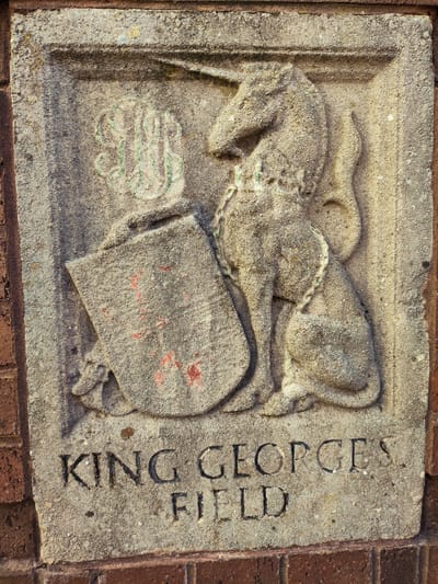 The image shows a stone relief carving affixed to a brick wall. The carving depicts a unicorn sitting on its hindquarters, facing left, with a chain around its neck. To the left of the unicorn is a shield, and above that is a monogrammed crest. The carving is weathered and shows signs of age and some staining, with traces of green paint on the monogram and red paint on the shield. Below the carving, carved into the stone, are the words "KING GEORGES FIELD". The relief is installed on the exterior of a building, as indicated by the surrounding brickwork. This stone relief is located at the entrance to King George's Field in Maghull, United Kingdom.