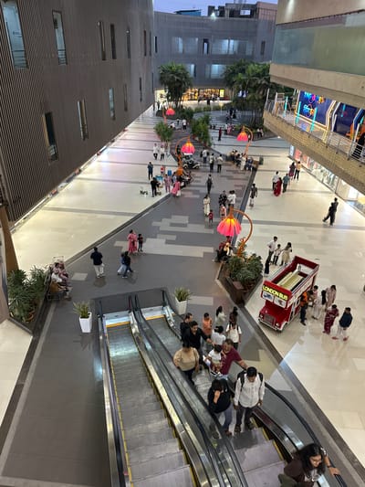 The image depicts a high-angle view of an outdoor mall area in Kalyan-Dombivli, India. People are walking and congregating in the mall. An escalator leads from one level to another. There is a small, red "London Bus" attraction. The architecture includes modern buildings with linear facade elements and some tropical greenery. The lighting suggests it is either dusk or an evening scene.