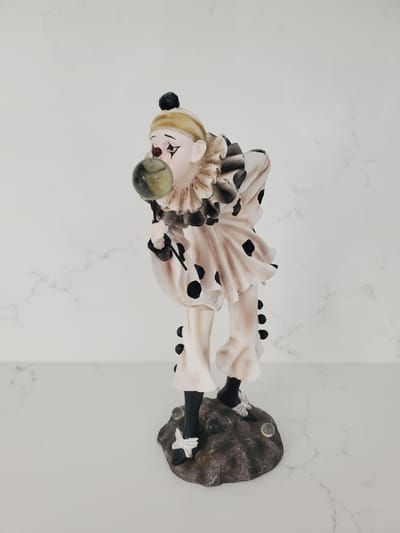 The image shows a ceramic figurine of a clown on a rock base. The clown is wearing a white outfit with black polka dots and a ruffled collar. The clown is holding a green and yellow marble up to its mouth, as if blowing a bubble. The background is a white marble countertop and wall. The scene is indoors and appears to be a still life or product photograph. There are no people or animals present in the image, and the location in Kitchener, Canada is not specified.