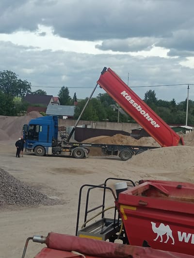 The image shows a blue MAN truck with a red Kässbohrer tipper semi-trailer in Яльгелево, Russia. The tipper is raised and is unloading sand. Two men are standing near the truck cab, engaged in conversation. In the foreground, there is a portion of another red trailer with a white camel logo. Gravel is piled to the left of the truck and sand piles surround the trailer. In the background are buildings and trees. The sky is overcast.