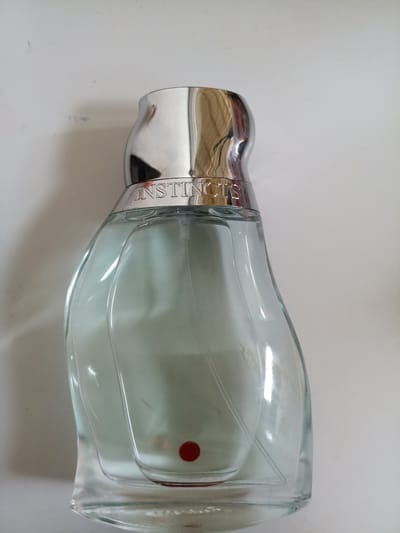 A clear glass perfume bottle with a silver cap, labeled "INSTINCTS," is placed on a white surface, possibly a table or countertop. The bottle contains a light green liquid. A small red circular sticker is visible on the lower front portion of the bottle. The background is a plain white wall with some faint smudges. The lighting is bright, casting soft shadows. The setting is an indoor environment in the city of Bishkek (formerly Frunze), Kyrgyzstan.