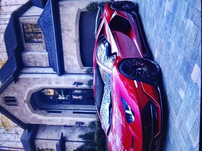 The image shows a red sports car parked on a paved driveway in front of a large, stone-built house with dark roofing. The car is positioned at an angle, with its side and front visible. The house features an arched entryway and several windows. The driveway is made of light-colored rectangular pavers. The overall scene suggests a luxurious residential setting. There are no people visible in the image. The image quality is somewhat grainy. The provided crops highlight details of the car's wheels and bodywork, as well as portions of the house's architecture and the driveway. It is not possible to determine the specific location of Safonovo, Russia from this image.