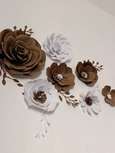 The image shows a collection of handmade paper flowers and leaves arranged on a white surface. There are seven flowers in total, with varying sizes and colors. Five of the flowers are a deep brown color, and two are white. The brown flowers have intricate centers, some with white accents, while the white flowers have brown centers. Several brown and white paper leaves and branches are interspersed among the flowers, creating a decorative arrangement. The lighting suggests an indoor setting, possibly a studio or home, with a soft light casting subtle shadows. No people are visible, and the focus is solely on the crafted floral display. The background is a plain white, which makes the colors and textures of the paper flowers stand out. The overall impression is one of a carefully arranged decorative piece.