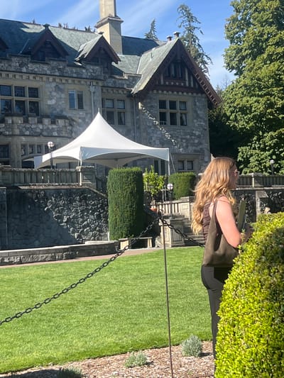 In the city of Langford, Canada, a woman with long blonde hair and sunglasses stands in front of a stone mansion. She is wearing a brown top and black pants, and is holding a green bag and a document. A white tent is set up on the lawn in front of the mansion. The mansion is a large, stone building with a dark roof and several windows. The lawn is green and well-manicured, with a chain link fence running across it. Tall trees are visible in the background. The sky is blue and clear. The scene appears to be a sunny day.