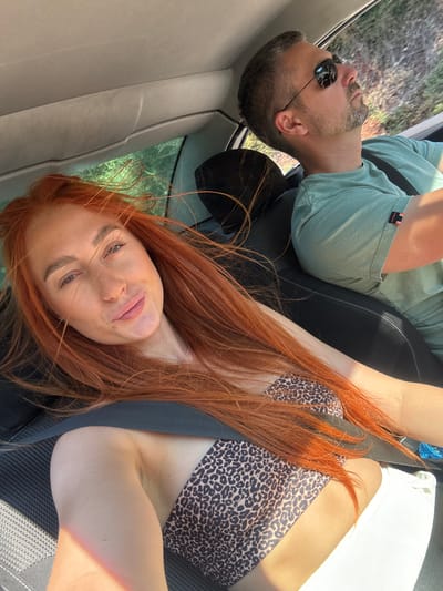 The image captures two people in a car. A man with short brown hair and a beard, wearing sunglasses and a green t-shirt, is seated in the driver's seat. He is looking upwards. Next to him, a woman with long, bright red hair is leaning towards the camera, her face slightly tilted and eyes looking at the viewer. She is wearing a leopard-print bikini top. The scene appears to be daytime, with sunlight streaming into the car, casting shadows and highlighting the subjects. The background visible through the car window suggests a natural, possibly rural or forested environment, consistent with the provided location context of Medinski Krš, Montenegro. The mood seems relaxed and informal, possibly during a road trip or outing. The visible text on the man's t-shirt is a small, orange and black label with white lettering, partially obscured.