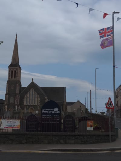 The image captures a street view in Magherafelt, United Kingdom, featuring a Presbyterian church with a prominent spire. Several flags are visible: a Union Jack and a purple flag with an emblem. Bunting stretches across the sky. A street sign with two stick figures indicates a pedestrian crossing. The church is flanked by a stone wall and a fence, behind which are gravestones and a banner advertising a "Tea & Coffee" event. The environment appears overcast with a hint of blue sky. No individuals are clearly depicted or engaged in interaction. The scene is static and likely captured during daylight hours.