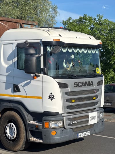 This is a photograph of a white Scania truck parked on a street in Dobieżyn, Poland. The truck has a prominent "SCANIA" logo on the front grille, and a "G 410" model designation. A white sign reading "ODPADY" is attached to the grille, indicating waste collection. The truck's license plate is "PKS 34229". A grey Renault Clio is partially visible behind the truck. Trees and a blue sky are visible in the background. The truck appears to be a utility vehicle, possibly for refuse collection, as suggested by the "ODPADY" sign. The scene is likely captured during daylight hours on a clear day.