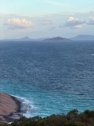 This image depicts a coastal landscape in the city of West Beach, Australia. The foreground features dense, low-lying green and brown coastal vegetation on the lower right, and a large, smooth, light brown and grey granite rock outcrop on the lower left, sloping towards the water. Turquoise ocean water with breaking white foam meets the rock and extends horizontally. The water transitions to a darker blue in the midground, with visible wave texture. A small, dark-colored boat with a white wake is observed traveling from right to left in the mid-right portion of the ocean. In the background, several islands and distant landmasses rise from the dark blue water, appearing as dark brown/green and blue-grey silhouettes against the horizon. The sky is a pale blue with scattered white cumulus clouds. No individuals are visible in the scene.