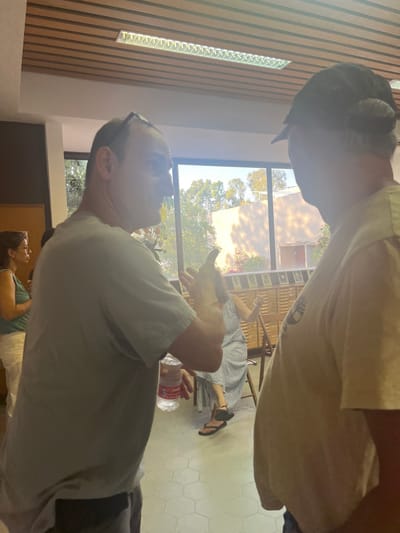 This image depicts an indoor scene within a building located in the Brener Regional Council, Israel, likely a community hall or meeting room, during daytime.
**People:**
Four individuals are visible.
1. In the foreground, a male with short dark hair and sunglasses perched on his head, wearing a light grey t-shirt and dark shorts, stands in profile facing right. He holds a clear plastic water bottle with a red label in his right hand and gestures with his left hand, palm open, mid-conversation.
2. To his right and partially obscuring the view, a second male with grey hair visible under a dark baseball cap, wearing a light-colored t-shirt, stands with his back mostly to the viewer, facing left towards the first male, engaging in conversation.
3. In the mid-ground to the left, a female with dark hair, wearing a light-colored sleeveless top and white trousers, is partially visible, standing and facing left.
4. In the mid-ground center-right, visible through the space between the two foreground males, a female with dark hair, wearing a light-colored, possibly patterned, long dress and dark flip-flops, is seated on a light wooden chair. She faces slightly left and gestures with both hands raised, palms open, appearing engaged in a discussion.
**Objects and Environment:**
The room features a wooden-slatted ceiling with recessed linear fluorescent light fixtures. The floor is covered in light-colored hexagonal tiles. Large windows span the background, revealing an outdoor scene with lush green trees and foliage, as well as the light-colored exterior of another building with a sloped roof. A wooden display cabinet or shelving unit, containing multiple small framed items (likely photographs or plaques), is situated along the wall beneath the windows, behind the seated female. The room is brightly illuminated by a combination of natural light from the windows and artificial overhead lighting.