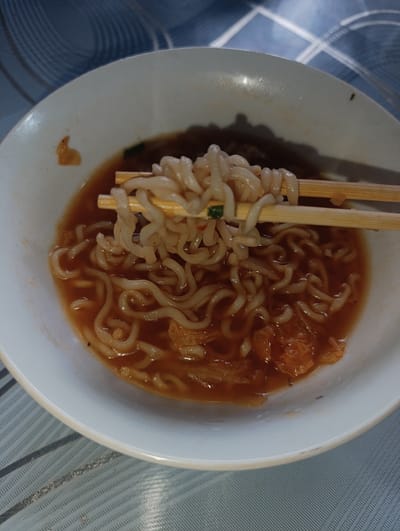 A close-up, overhead shot captures a white bowl filled with ramen noodles and broth, resting on a striped placemat. Chopsticks are lifting a portion of the noodles from the bowl. The broth is a reddish-brown color, and some orange pieces, possibly kimchi or vegetables, are visible within the bowl. The background is blurred, showing a hint of a patterned surface. The image does not contain information about the city of Novopokrovka, Kyrgyzstan.