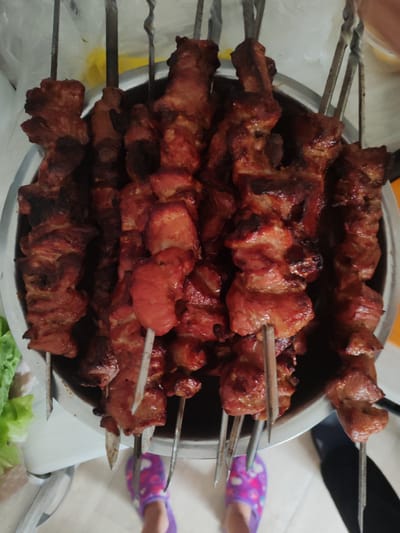 A close-up, overhead shot showcases a bowl filled with several skewers of grilled meat, likely pork, given its reddish-brown hue and visible fat marbling. The meat pieces are roughly cube-shaped and appear to be well-cooked, with some charred edges indicating grilling. Metal skewers pierce through the meat, with some extending out of the bowl. The bowl itself is metallic and circular. To the left of the bowl, a head of lettuce is partially visible, suggesting a potential side dish or garnish. In the lower portion of the image, two feet are visible, one wearing a purple slipper with a flower pattern. The background is blurred but appears to be an indoor setting, possibly a kitchen or dining area, with hints of a plastic bag and a yellow object. The lighting is overhead and slightly warm.