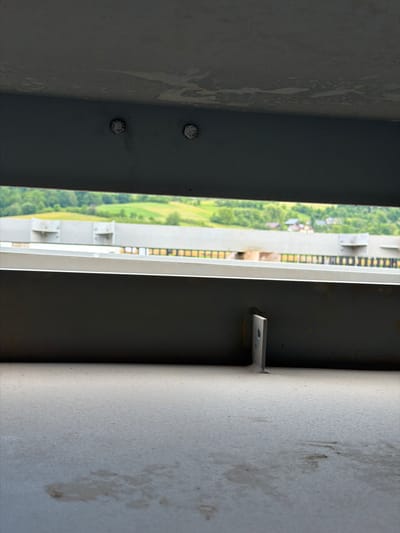 This image captures a close-up view of a weathered metal structure, possibly part of a bridge or industrial building, with a scenic rural landscape visible in the background. The foreground is dominated by a rough, light-colored concrete surface with faint stains. Above this, a dark gray metal beam runs horizontally, showing signs of wear and tear, including chipped paint and possibly some rust. Two large, hexagonal bolts are prominently visible on the underside of this beam.

Further back, another dark gray metal beam is present, and on its surface, a small, L-shaped metal bracket is attached. Through the gap between these two beams, a wider view reveals a distant landscape characterized by rolling green hills dotted with trees and a few scattered houses with red roofs. A lighter-colored structure, possibly another bridge or elevated walkway with a black railing, spans across the middle ground, partially obscuring the view of the landscape. The overall impression is one of an outdoor, man-made environment contrasted with a natural, pastoral setting. The image appears to be taken from a low vantage point, looking upwards and outwards. There are no people or active elements depicted in the scene.

The location is identified as Jodłownik, Poland.