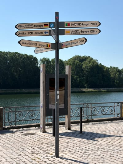 The image depicts a directional signpost located in the city of Charnay-lès-Mâcon, France. The signpost has multiple arms pointing in various directions, indicating distances and destinations in kilometers. These destinations include NEUSTADT in Germany, PORI in Finland, OVERIJSE in Belgium, EGER in Hungary, SANTO TIRSO in Portugal, ALCAZAR de SAN JUAN in Spain, and MACON in the United States. The signpost is situated on a paved area beside a body of water, likely a river or lake, bordered by a decorative metal railing. In the background, dense green trees line the opposite bank of the water. A tall, dark vertical pole is prominently visible in the foreground, likely part of the signpost structure. A wooden post with circular emblems is also present, along with a display board containing text and images, possibly an informational exhibit. The sky is clear and blue.