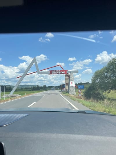 The image shows a view from inside a vehicle looking out at a road leading towards a large archway that marks the entrance to the county of Brasov, Romania. The archway has a modern, abstract design with a prominent white triangular structure. Red beams extend from the top of the arch. A stone pillar supporting the archway displays the coat of arms of the Brasov County Council and text reading "Consiliul Judetean Brasov." A blue road sign to the right of the archway indicates "Județul Brașov" with an emblem and a warning for uneven road surface. The road is paved and has white lane markings. In the background, there are green fields, trees, rolling hills, and a partly cloudy blue sky. Several vehicles, including trucks and cars, are visible on the road in the distance. The foreground shows the dashboard of the vehicle from which the photo was taken. The location is identified as the entrance to the county of Brasov, in Ucea de Jos, Romania.